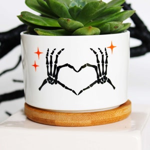 May include: White ceramic planter with a black skeleton hand design making a heart shape. The planter has orange stars and is filled with a green succulent plant. The planter is sitting on a wooden base.