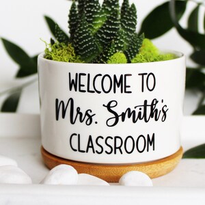 May include: A white ceramic pot with the text "WELCOME TO Mrs. Smith's CLASSROOM" in black lettering. The pot contains a succulent arrangement with green plants and moss, sitting on a bamboo saucer. Decorative white stones are in the foreground.