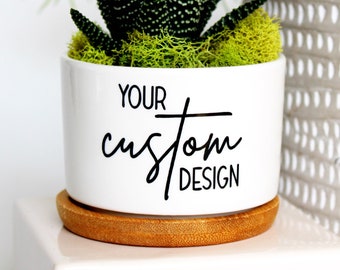 May include: A white ceramic plant pot with the text "YOUR CUSTOM DESIGN" in black script. It contains a succulent plant with green moss and rests on a small wooden saucer. Another plant in a grey textured pot is in the background.
