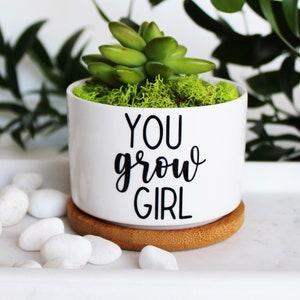Puede incluir: Maceta de cer&aacute;mica blanca con una planta suculenta y el texto "You grow girl" en letras negras. La maceta tiene una base de bamb&uacute;.