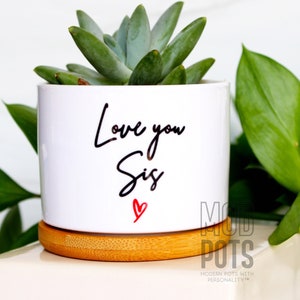 May include: White ceramic planter with a bamboo base. The planter has a handwritten message that says "Love you Sis" with a red heart.
