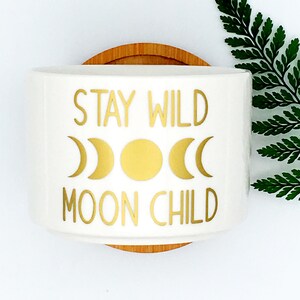 Puede incluir: Maceta de cer&aacute;mica blanca con una inscripci&oacute;n dorada que dice "Stay Wild Moon Child". La inscripci&oacute;n est&aacute; rodeada de tres lunas crecientes doradas y un sol dorado.