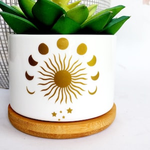 May include: White ceramic planter with a gold sun and moon design. The planter has a bamboo base and is decorated with a gold sun and crescent moons. The planter is perfect for small plants or succulents.