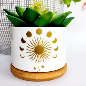 May include: A white cylindrical ceramic planter featuring a gold metallic sun graphic surrounded by a lunar phase cycle and small stars. The planter sits on a circular bamboo wood saucer and contains a green artificial succulent plant.