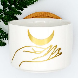 May include: White ceramic candle jar with a gold crescent moon design held in a stylized hand. The jar has a bamboo lid.
