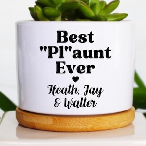 May include: A white ceramic planter with the text "Best "PI"aunt Ever" in black, holding a small green succulent. The planter sits on a wooden coaster atop a white square base, with green leaves in the background.