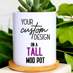 Peut inclure: Un pot en céramique blanche avec une base en bois. Le pot porte l'inscription "YOUR custom DESIGN on a TALL MOD POT" en lettres noires et violettes. Une petite plante succulente pousse dans le pot.
