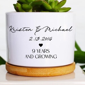 May include: A white ceramic planter with a succulent plant, resting on a wooden coaster and a white square base. The planter is inscribed with the names "Kristen & Michael", the date "2.13.2016", a heart symbol, and the phrase "9 YEARS AND GROWING".