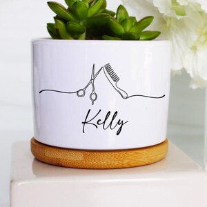 May include: White ceramic planter with a bamboo base, featuring a black line drawing of scissors and a comb, with the name "Kelly" written below. A small succulent plant is growing in the planter.