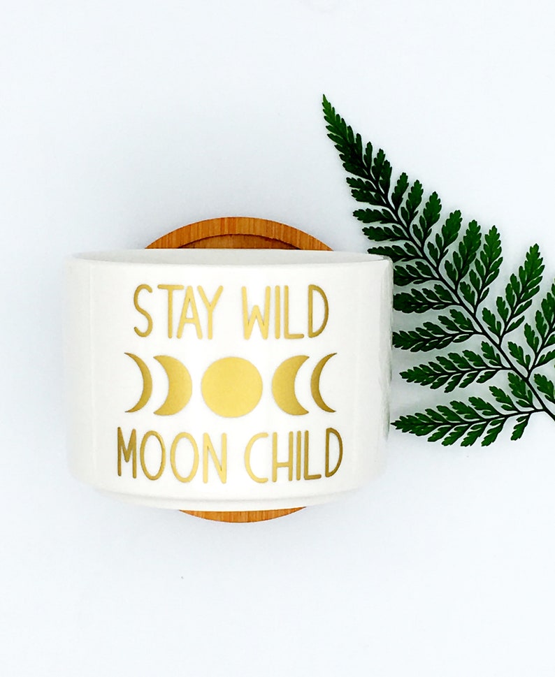 Puede incluir: Maceta de cer&aacute;mica blanca con una inscripci&oacute;n dorada que dice "Stay Wild Moon Child". La inscripci&oacute;n est&aacute; rodeada de tres lunas crecientes doradas y un sol dorado.