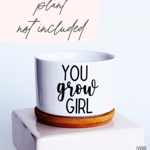 Puede incluir: Maceta de cer&aacute;mica blanca con base de madera. La maceta tiene texto negro que dice "You grow girl".