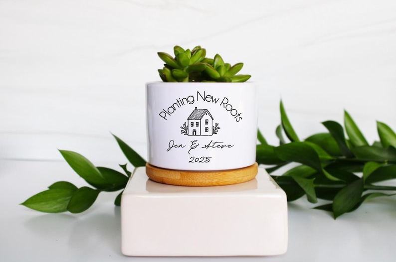 May include: A white ceramic planter with the text "Planting New Roots" and a house graphic, personalized with names and the year 2025. The planter sits on a wooden base and contains a green succulent plant. The planter is on a white stand.