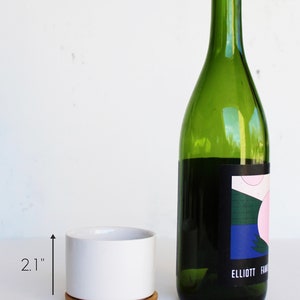 Puede incluir: Una maceta de cer&aacute;mica blanca con una base de madera, que mide 2,1 pulgadas de alto y 3,15 pulgadas de ancho, junto a una botella de vino verde est&aacute;ndar con una etiqueta colorida. La etiqueta presenta un dise&ntilde;o geom&eacute;trico con colores rosa, azul y verde. La etiqueta tambi&eacute;n incluye el texto "ELLIOTT FAM".