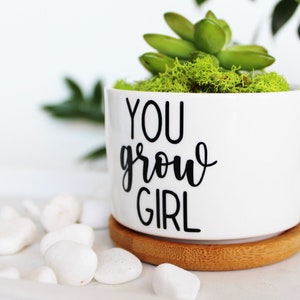 Puede incluir: Maceta de cer&aacute;mica blanca con el texto "YOU grow GIRL" en letras negras. La maceta est&aacute; llena de musgo verde y una peque&ntilde;a planta suculenta. La maceta est&aacute; sobre un posavasos de madera.