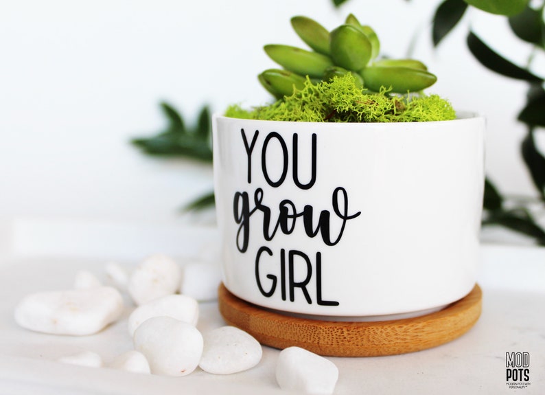 Puede incluir: Maceta de cer&aacute;mica blanca con el texto "YOU grow GIRL" en letras negras. La maceta est&aacute; llena de musgo verde y una peque&ntilde;a planta suculenta. La maceta est&aacute; sobre un posavasos de madera.