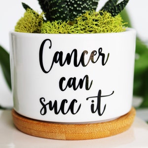 May include: White ceramic pot with the text "Cancer can succ it" in black script. The pot contains a succulent plant with green and yellow moss. The pot sits on a round, light brown bamboo coaster.