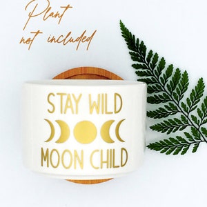 Puede incluir: Maceta de cer&aacute;mica blanca con una inscripci&oacute;n dorada que dice "Stay Wild Moon Child". La inscripci&oacute;n incluye un sol estilizado y fases lunares. La maceta est&aacute; sobre una base de madera.