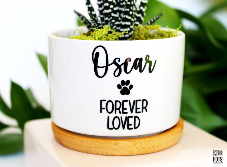 May include: White ceramic planter with a bamboo base. The planter has black lettering that reads "Oscar Forever Loved" with a paw print symbol.
