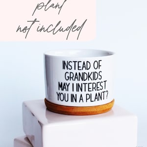 May include: White ceramic plant pot with the text "INSTEAD OF GRANDKIDS MAY I INTEREST YOU IN A PLANT?" in black. The pot sits on a round, natural wood coaster. The words "plant not included" are above the pot.