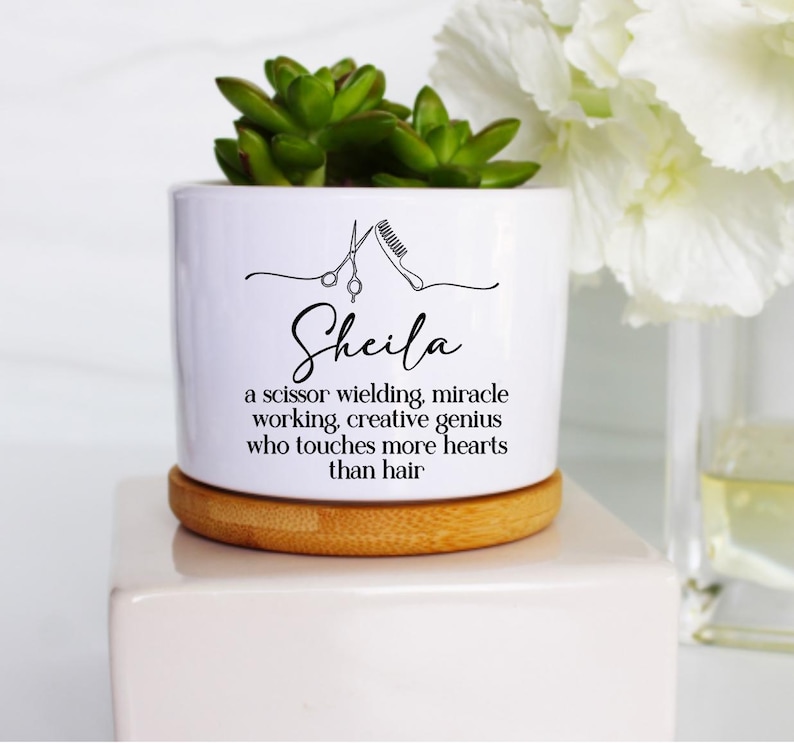 May include: A white ceramic planter with a bamboo base containing a green succulent. The planter features a black line drawing of scissors and a comb, the name Sheila, and the text: a scissor wielding, miracle working, creative genius who touches more hearts than hair.