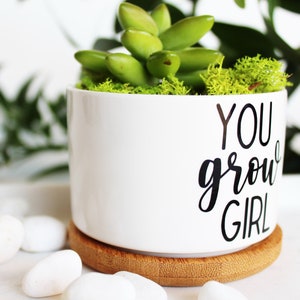 Puede incluir: Maceta de cer&aacute;mica blanca con base de bamb&uacute;. La maceta tiene el texto "YOU grow GIRL" en letras negras. La maceta est&aacute; llena de suculentas verdes y musgo.