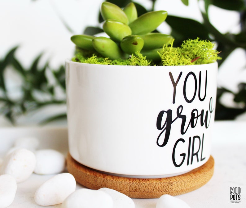 Puede incluir: Maceta de cer&aacute;mica blanca con base de bamb&uacute;. La maceta tiene el texto "YOU grow GIRL" en letras negras. La maceta est&aacute; llena de suculentas verdes y musgo.