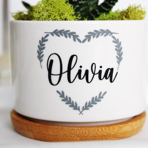 May include: White ceramic planter with a gray heart-shaped wreath and the name "Olivia" in black lettering. The planter has a wooden base.