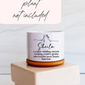 May include: A white cylindrical ceramic planter with a bamboo base. The front features a black line drawing of scissors and a comb above the name Sheila and the text: a scissor wielding, miracle working, creative genius who touches more hearts than hair.
