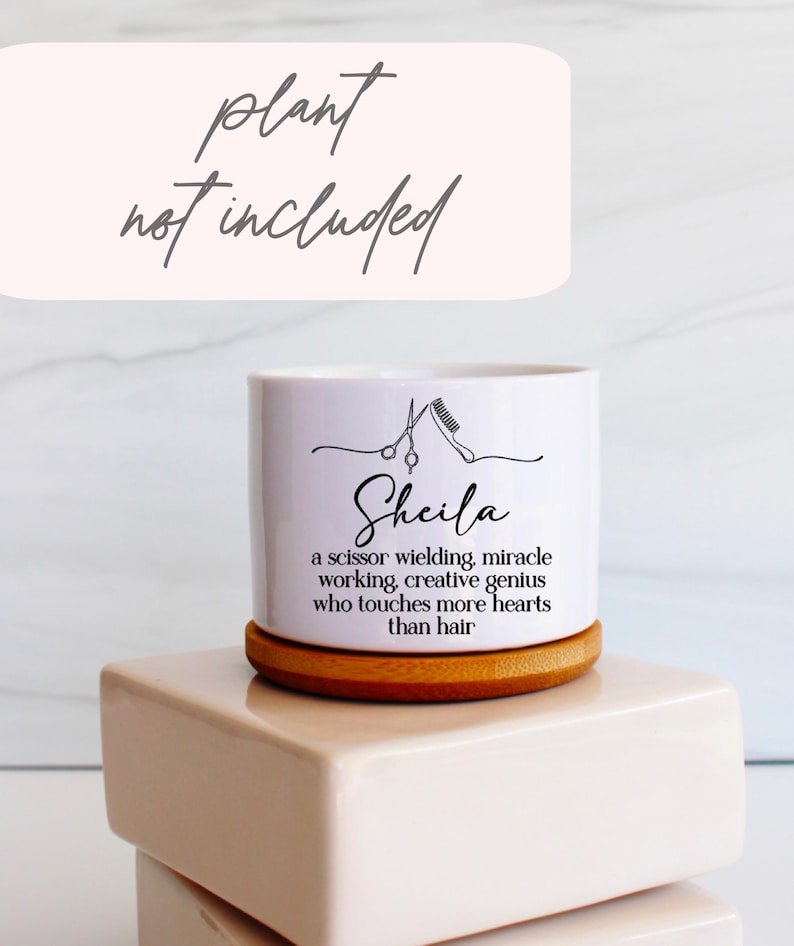 May include: A white cylindrical ceramic planter with a bamboo base. The front features a black line drawing of scissors and a comb above the name Sheila and the text: a scissor wielding, miracle working, creative genius who touches more hearts than hair.