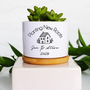 K&ouml;nnte beinhalten: Ein wei&szlig;er Keramik-Pflanztopf mit einer Sukkulentenpflanze, der auf einem Holzuntersetzer und einem wei&szlig;en Block steht. Der Topf tr&auml;gt den Text "Planting New Roots" und die Namen "Jen & Steve" mit dem Jahr "2025".
