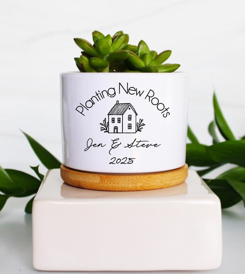 K&ouml;nnte beinhalten: Ein wei&szlig;er Keramik-Pflanztopf mit einer Sukkulentenpflanze, der auf einem Holzuntersetzer und einem wei&szlig;en Block steht. Der Topf tr&auml;gt den Text "Planting New Roots" und die Namen "Jen & Steve" mit dem Jahr "2025".