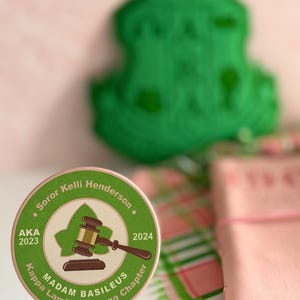 May include: A round, brown and green paper coaster with the text "Soror Kelli Henderson", "AKA 2023", "2024", "Madam Basileus", "Kappa Lambda Omega Chapter" and a gavel and green leaf design.
