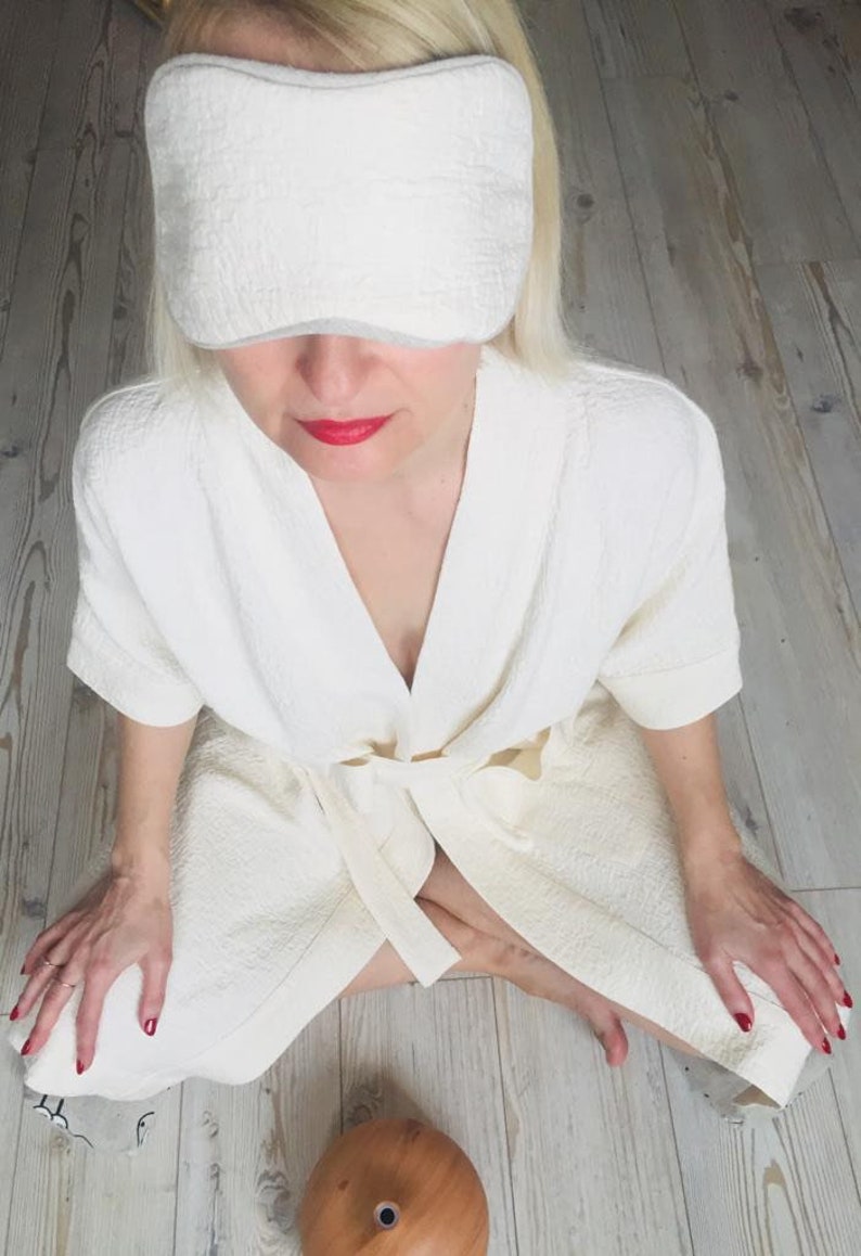May include: A person wearing a white robe and a white eye mask sits on a wooden floor with a wooden essential oil diffuser in front of them.