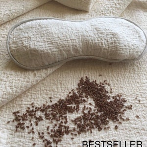 May include: A white eye mask made of textured fabric, with a light gray trim. The eye mask is lying on a white textured fabric surface. A scattering of brown flax seeds are on the surface below the eye mask. The text "BESTSELLER" is printed in black on the fabric.