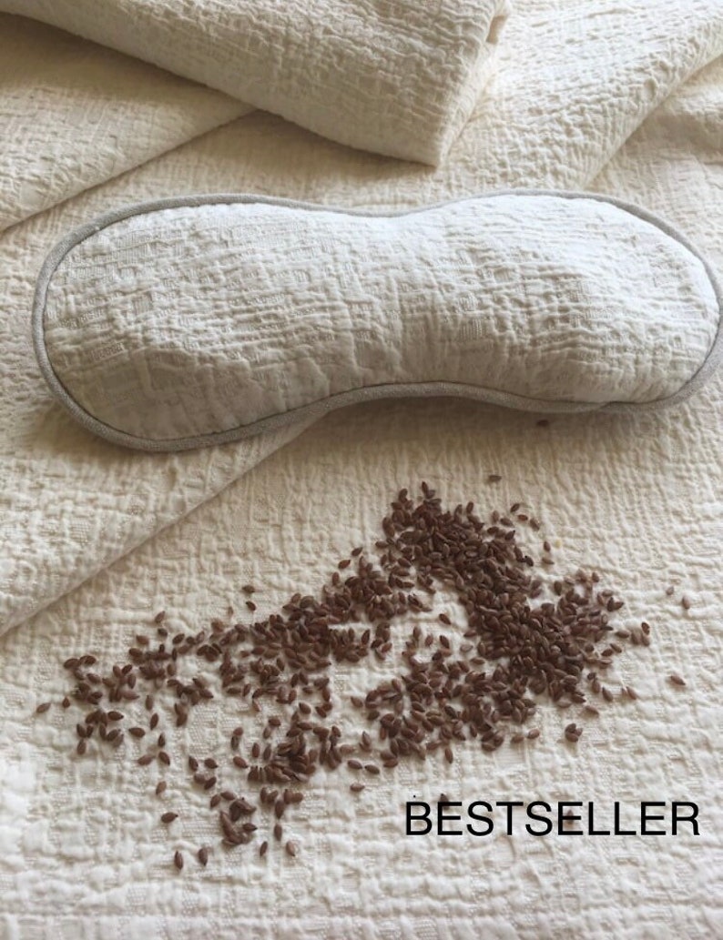 May include: A white eye mask made of textured fabric, with a light gray trim. The eye mask is lying on a white textured fabric surface. A scattering of brown flax seeds are on the surface below the eye mask. The text "BESTSELLER" is printed in black on the fabric.