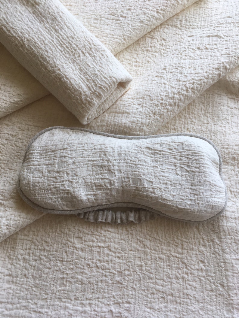 May include: A white eye mask with a ruffled edge and a textured fabric. The mask is lying on a white textured fabric.