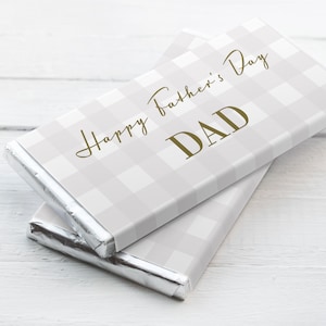 Könnte beinhalten: Zwei Schokoladentafeln, verpackt in Silberfolie mit einem weißen und grauen Gingham-Muster. Die obere Tafel hat den Text "Happy Father's Day DAD" in Gold gedruckt.