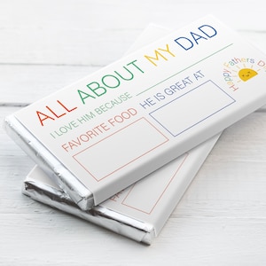 Könnte beinhalten: Eine weiße Schokoladentafel mit einem bunten Design, auf dem "All About My Dad" steht. Es gibt zwei leere Felder zum Eintragen, eines für "Ich liebe ihn, weil" und das andere für "Lieblingsessen, in dem er großartig ist". Am unteren Rand der Tafel befindet sich eine kleine Sonne mit dem Text "Happy Fathers Day".