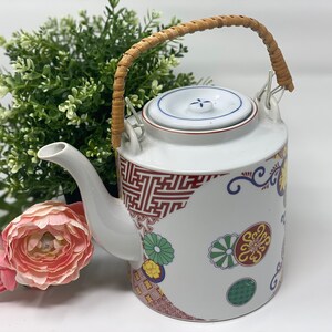 May include: White ceramic teapot with a brown wicker handle and a lid with a blue and white floral design. The teapot is decorated with red, blue, green, and yellow floral patterns.