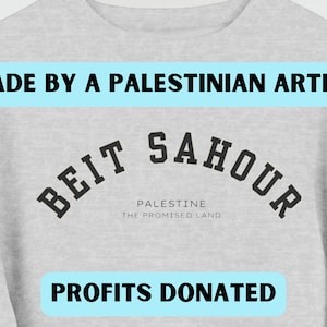 Può includere: Felpa grigia con testo nero che recita "Beit Sahour" in un arco. Il testo "Palestine The Promised Land" è sotto l'arco. Il testo "Made by a Palestinian Artist" è in un riquadro blu nella parte superiore dell'immagine. Il testo "Profits Donated" è in un riquadro blu nella parte inferiore dell'immagine.