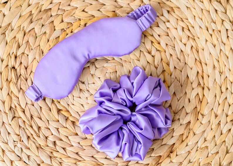 May include: A lavender satin sleep mask and a lavender satin scrunchie on a woven straw surface.