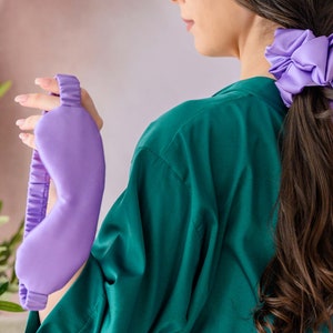 May include: A purple satin sleep mask with an elastic band. The mask is held by a hand wearing a green shirt. A purple satin scrunchie is in the hair of the person holding the mask.