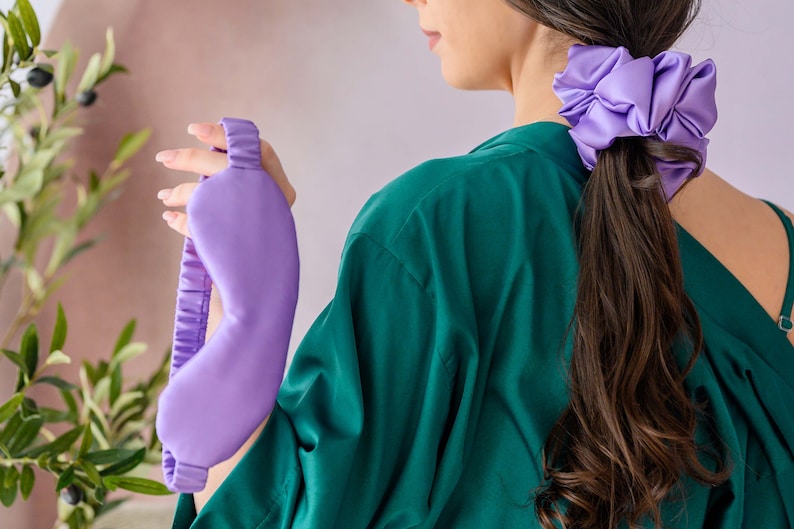 May include: A purple satin sleep mask with an elastic band. The mask is held by a hand wearing a green shirt. A purple satin scrunchie is in the hair of the person holding the mask.