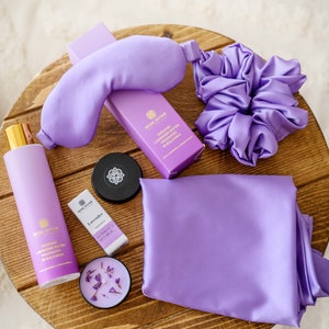 May include: A lavender-themed self-care gift set with a sleep mask, hair scrunchie, candle, essential oil, and lavender water. The products are all in shades of purple and are arranged on a wooden surface.