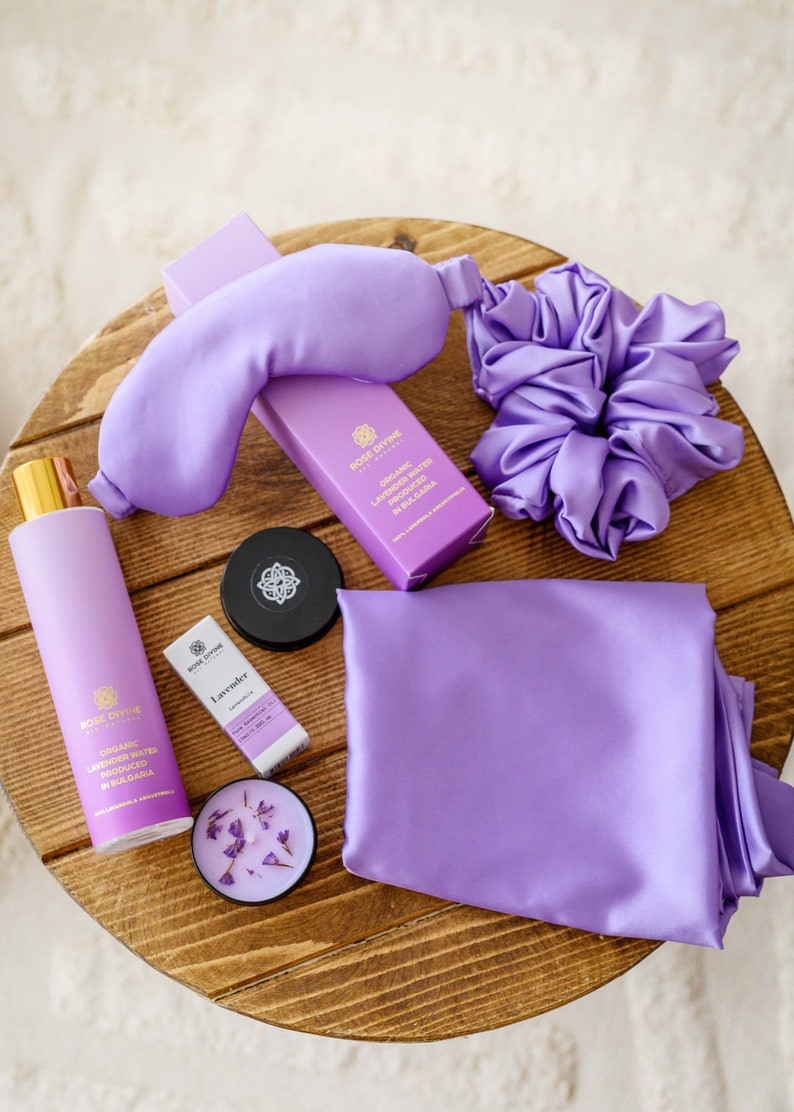 May include: A lavender-themed self-care gift set with a sleep mask, hair scrunchie, candle, essential oil, and lavender water. The products are all in shades of purple and are arranged on a wooden surface.
