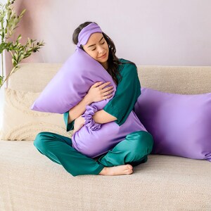 May include: A person wearing a green and purple satin pajama set and a purple sleep mask is sitting on a couch and holding a purple pillow.