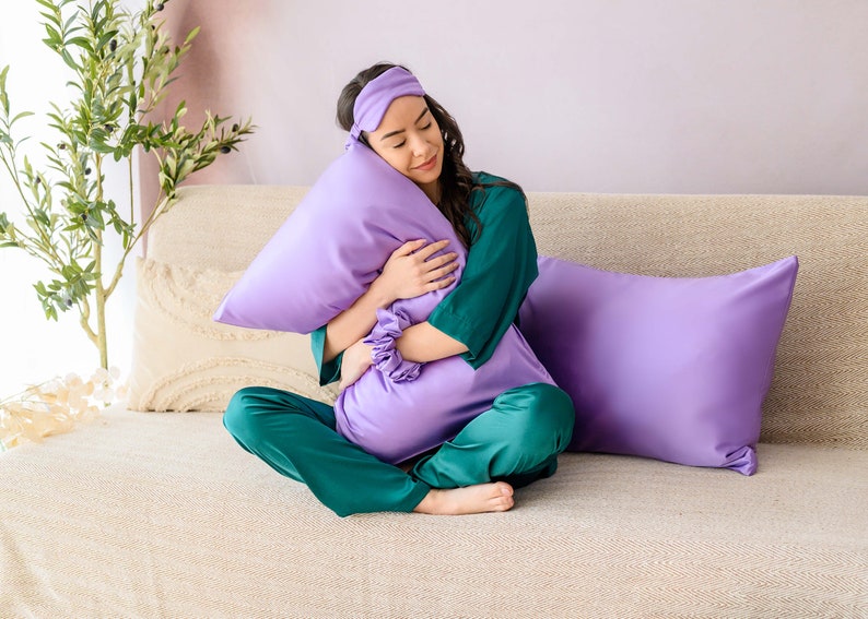 May include: A person wearing a green and purple satin pajama set and a purple sleep mask is sitting on a couch and holding a purple pillow.