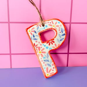 May include: A ceramic letter "P" ornament with a floral design in red, blue, and yellow. The letter has a white background and a red border. It hangs from a brown string against a pink and purple backdrop.