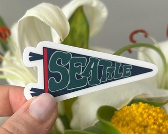 May include: A white pennant with a blue and green design that says "SEATTLE" in a retro font. The pennant is held in a hand against a background of white flowers.