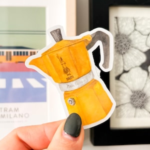 May include: A yellow sticker of a stovetop espresso maker, held in front of framed artwork. The sticker has a watercolor effect and features the brand name. The background includes a framed picture of a tram and a black and white floral print.